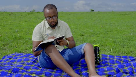 A-black-man-sits-on-a-blanket-in-the-grass-and-takes-notes-in-a-book