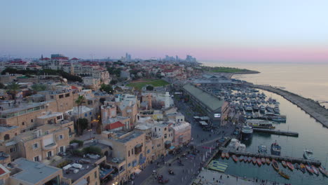 Old-city-of-Jaffa-and-Jaffa-port-at-sunset-with-lots-of-families-visiting-restaurants,-shops-and-bars-in-the-port---parallax-drone-shot