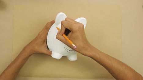 Drawing-happy-face-on-white-ceramic-piggy-bank-on-brown-paper-at-home