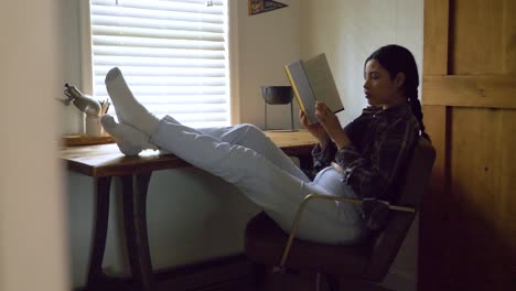 Focused-Puerto-Rican-female-reading-captivating-story-with-feet-raised-resting-on-desk