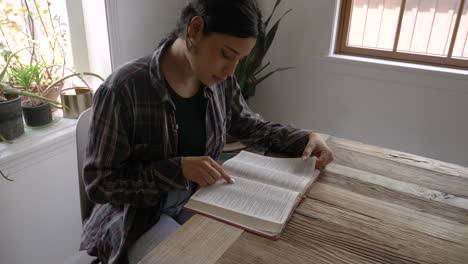 Bequemes-Puerto-ricanisches-Mädchen,-Das-Die-Heilige-Bibel-Liest-Und-Sich-Am-Holztisch-Mit-Hellem-Fenster-Entspannt