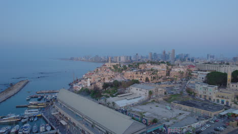 Wide-parallax-view-drone-shot-of-Old-city-of-Jaffa-and-Jaffa-port-at-sunset-with-lots-of-families-visiting-restaurants,-shops-and-bars-in-the-port---tel-aviv-city-at-the-background