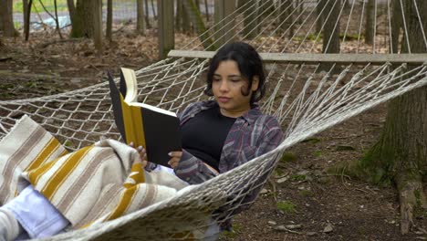 Comfortable-young-Puerto-Rican-woman-reading-book-swinging-in-woodland-hammock