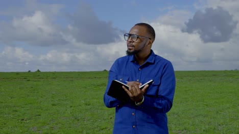 A-black-man-stands-in-a-meadow-counting-something-and-taking-notes-in-a-book