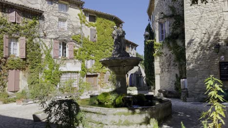 Picturesque-romantic-small-square-with-old-stone-houses-with-ivy-and-a-small-fountain-with-a-figure-in-the-warm-south-of-France