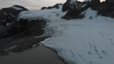 Aerial-view-of-the-glacial-lake-of-Claridenfirn-glacier-in-Uri,-Swizerland-at-dusk-with-a-pan-up-view-to-the-glowing-sky-behind-the-alpine-peaks-from-iceberg-filled-water