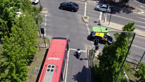 A-Red-Bus-Waits-in-Santiago-de-Chile-Streets-Traffic-Lights,-Aerial-Drone,-Taxi-and-Cars-Drive-along-Las-Condes-Neighborhood