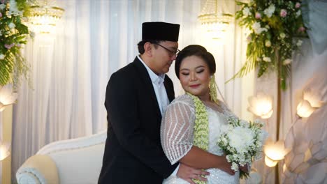 Auf-Einer-Indonesischen-Hochzeitsfeier-Umarmt-Der-Bräutigam-Die-Braut-Von-Hinten,-Sie-Tragen-Traditionelle-Indonesische-Hochzeitskleider-Und-Tragen-Einen-Blumenstrauß