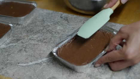 Chocolate-paste-spread-over-brownie-in-aluminum-container-using-baking-spoon,-filmed-as-close-up-in-slow-motion-style