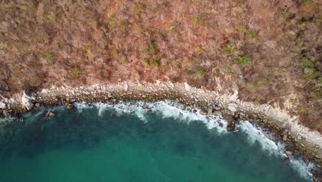 Aerial-top-down-shot-showing-waves-of-Pacific-Ocean-reaching-rocky-coastline-in-Huatulco,-México