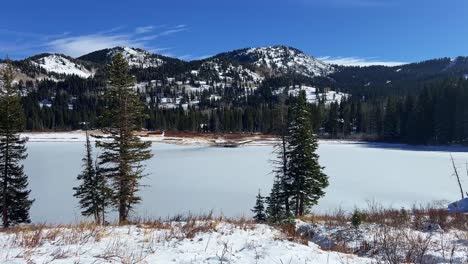 Tilt-up-handheld-extreme-wide-landscape-nature-shot-of-the-frozen-Silver-Lake-surrounded-by-pine-trees-in-the-famous-ski-town-of-Brighton,-Utah-on-a-sunny-warm-winter-day