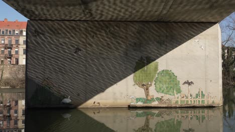 Graffiti-art-at-the-wall-near-the-reflective-water-surface,-and-concrete-wall-holding-the-bridge-structure-above-displays-an-artsy-message,-location-Sarreguemines,-France