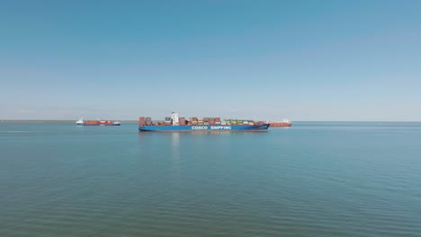 An-aerial-view-captures-a-loaded-container-ship-heading-out-into-the-Gulf-of-Mexico,-while-a-pleasure-boat-passes-by-at-the-east-end-of-Galveston-Island-under-clear-blue-skies-in-Galveston,-Texas