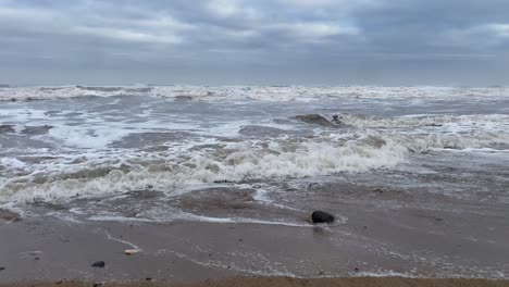 Low-angle-shot-of-rough-North-Sea-crashing-onto-sandy-beach-during-cloudy-day