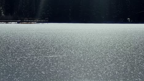 Tilt-up-slow-motion-handheld-shot-of-the-frozen-Silver-Lake-revealing-a-wooden-boardwalk-and-pine-trees-in-the-famous-ski-town-of-Brighton,-Utah-on-a-sunny-warm-winter-day
