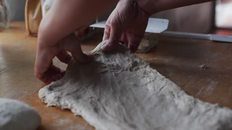 Fresh-dough-stretched-by-hand-over-on-wooden-tabletop,-filmed-in-close-up-handheld-slow-motion-style