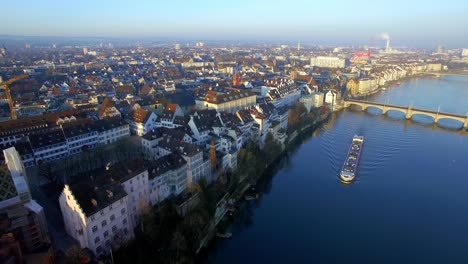 Embark-on-a-breathtaking-drone-journey-from-Basel's-skyline,-gliding-past-the-iconic-Minster,-and-unveiling-a-mesmerizing-sunrise-scene-with-a-cargo-ship-below