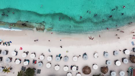 Die-Drohne-Fliegt-Von-Oben-Nach-Unten-über-Einem-Weißen-Strand-Mit-Sonnenschirmen-Und-Menschen,-Die-Im-Aquablauen-Ozean-Schwimmen,-In-Curaçao,-Luftaufnahmen-4k