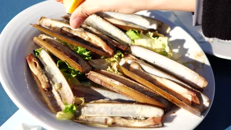 hand-putting-squeezed-lemon-on-grilled-sea-clams-on-white-plate-with-lettuce