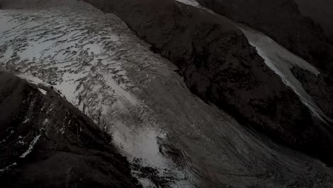 Aerial-flyover-over-Glacier-de-Vouasson-near-Arolla-in-Valais,-Switzerland-with-a-pan-up-view-from-the-icy-crevasses-to-the-colorful-sunset-glow-behind-the-layers-of-mountain-peaks