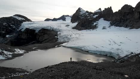 Aerial-flyover-over-a-hiker-overlooking-the-glacial-lake-and-ice-of-the-Claridenfirn-glacier-in-Uri,-Swizerland-with-a-view-of-the-alpine-peaks-and-icebergs-floating-in-the-water-during-sunset