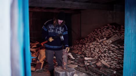 Man-Chopping-Wood-With-Axe-In-A-Barn-With-Pile-Of-Firewood