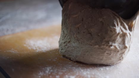 Soft-and-fluffy-batch-of-freshly-raised-dough-being-scraped-out-of-metal-bowl-onto-wooden-kitchen-tabletop,-filmed-as-closeup-slow-motion-shot