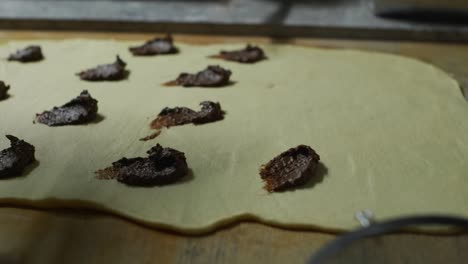Chocolate-paste-spooned-and-blotted-onto-freshly-rolled-white-dough-spread-over-wooden-kitchen-table-top,-filmed-as-close-up-slow-motion-shot