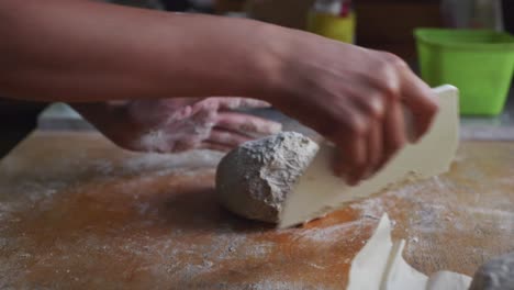 Ball-of-freshly-risen-dough-being-shaped-and-shuffled-by-hand-and-scraper-tool-on-wooden-kitchen-tabletop,-filmed-as-closeup-slow-motion-shot