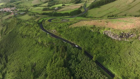 Drone-view-of-white-car-drives-on-small-road-in-green-scenery-at-Azores,-aerial