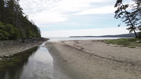 Calm-Aerial-Dolly-Shot-Over-a-Stream-Leading-Out-to-Coastal-Beach-with-Sand,-Sandpit,-Canada