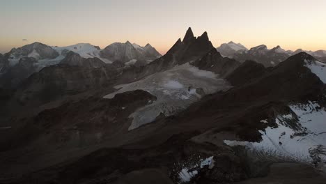 Aerial-flyover-over-Glacier-de-Vouasson-near-Arolla-in-Valais,-Switzerland-at-sunset-with-colorful-glow-behind-the-Aiguilles-Rouges,-Mont-Blanc-de-Cheillon,-Grand-Combin-and-other-peaks