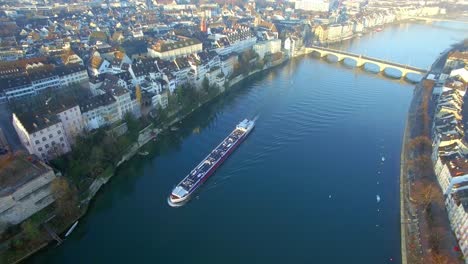 Stunning-sunrise-captured-by-a-drone:-a-cargo-ship-in-the-Rhine-river,-leaving-behind-the-bridge-after-emerging-from-it,-moving-forward-in-the-morning-light-of-Basel,-Switzerland