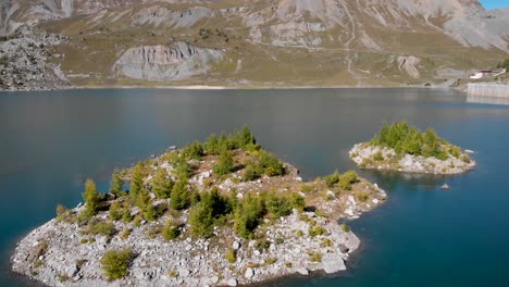Aerial-flyover-over-an-island-in-the-waters-of-Lac-de-Salanfe-in-Valais,-Switzerland-on-a-sunny-autumn-day-in-the-Swiss-Alps-with-a-view-of-surrounding-alpine-peaks,-cliffs-and-hydroelectric-dam