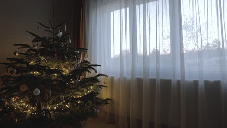Lighted-Christmas-Tree-Decor-Next-To-Window-With-Curtain-Through-Sunset