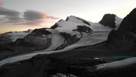 Sobrevuelo-Aéreo-Sobre-Un-Excursionista-Con-Vistas-A-Los-Glaciares-Allalin-Y-Hohlaub-Desde-El-Mirador-Cerca-De-Britattanihütte-En-Saas-fee,-Valais,-Suiza-Durante-Una-Colorida-Puesta-De-Sol-Con-Vistas-A-Los-Picos-Alpinos