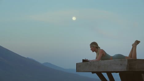 Female-traveler-journaling-while-laying-on-wooden-platform-with-full-moon,-Spiritual
