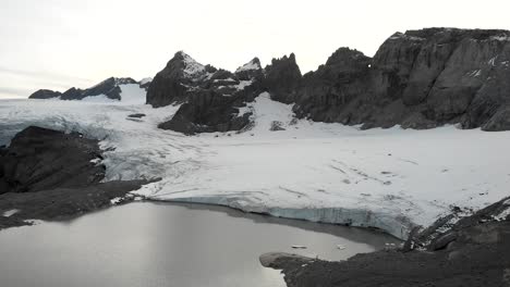 Eine-Sich-Drehende-Luftaufnahme-Des-Gletschersees-Des-Claridenfirn-Gletschers-In-Uri,-Schweiz-In-Der-Abenddämmerung-Mit-Dem-Leuchtenden-Himmel-Hinter-Den-Alpengipfeln-Und-Schwimmenden-Eisbergen-Im-Wasser