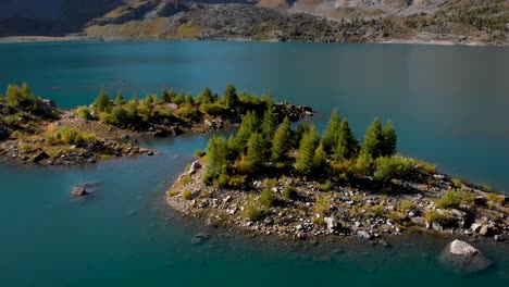 Aerial-flyover-over-an-island-in-the-waters-of-Lac-de-Salanfe-in-Valais,-Switzerland-on-a-sunny-autumn-day-in-the-Swiss-Alps-with-a-pan-down-view-from-the-surrounding-alpine-peaks-and-cliffs