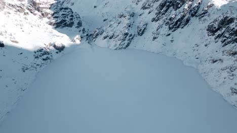 Aerial-of-frozen-alpine-lake-at-foot-of-rugged,-snowy-mountains,-Campagneda,-Italy