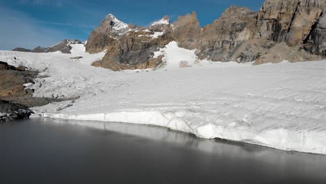 Sobrevuelo-Aéreo-Sobre-El-Lago-Glacial-Y-El-Hielo-Del-Glaciar-Claridenfirn-En-Uri,-Suiza,-En-Una-Mañana-Tranquila-Y-Pacífica-Con-Picos-Alpinos-Detrás-De-La-Pared-De-Hielo-Y-Su-Reflejo