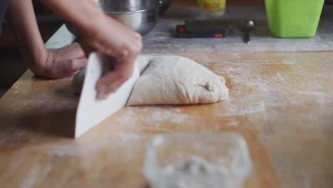 Freshly-kneaded-dough-being-cut-by-scraping-tool-on-wooden-kitchen-tabletop,-filmed-as-closeup-handheld-slow-motion-style
