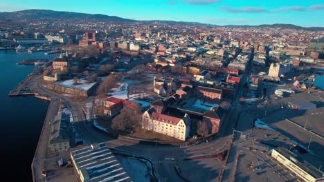 Oslo-Aerial-drone-at-waterfront-Central-train-station,-opera-and-Akershus-Fortress-along-the-seaside-fjord,-Norway-during-sunset