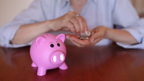 Woman's-Hand-Putting-Coins-On-The-Piggy-Bank-In-The-Wooden-Table