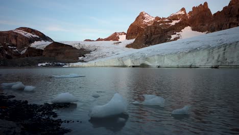 Peaceful-morning-scene-at-the-glacial-lake-next-to-Claridenfirn-glacier-in-Uri,-Swizerland-with-the-alpenglow-of-the-alpine-peaks-reflected-and-icebergs-next-to-the-shore