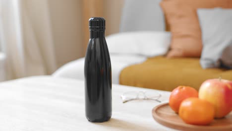 Frau-Stellt-Zu-Beginn-Des-Tages-Eine-Schwarze-Wasserflasche-Neben-Obst-Auf-Den-Tisch
