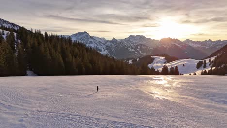 Ascending-drone-shot-showing-person-walking-on-snowy-slope-in-Swiss-Mountains-during-golden-sunset