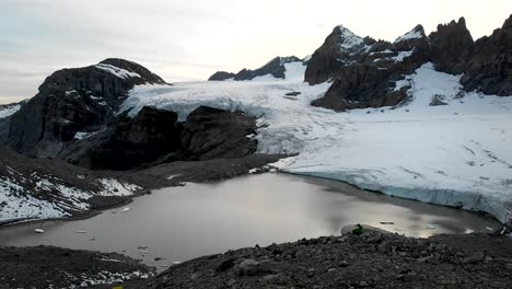 Aerial-flyover-over-a-hiker-enjoying-the-view-of-the-glacial-lake-of-Claridenfirn-glacier-in-Uri,-Swizerland-at-dusk-with-the-glowing-sky-behind-the-alpine-peaks-and-floating-icebergs-in-the-water