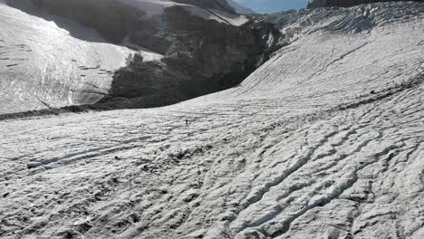 Sobrevuelo-Aéreo-Sobre-Un-Excursionista-Y-Montañero-Caminando-Sobre-El-Hielo-Y-Saltando-Sobre-Las-Grietas-Del-Glaciar-Allalin-Cerca-De-Saas-fee-En-Valais,-Suiza-En-Un-Día-Soleado-En-Los-Alpes-Suizos