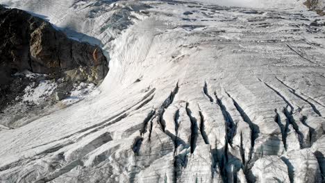 Aerial-flyover-over-a-glacier-near-Arolla-in-Valais,-Switzerland-with-a-pan-up-view-from-the-crevasses-in-the-glacier-ice-up-to-the-Aiguilles-Rouges-peaks-sunlit-by-the-early-morning-sunshine